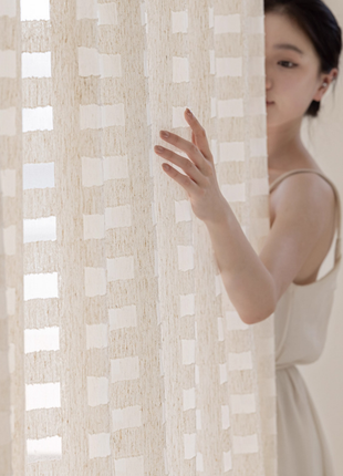 A woman in a white dress stands behind a beige curtain, her hand reaching out to the curtain.
