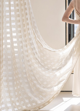 A woman in a white dress is opening a large, white, checkered curtain in a room with a window.