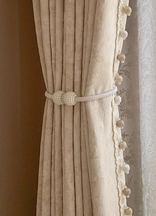A beige curtain with a decorative trim and a white knotted tie at the bottom, hanging in a room.