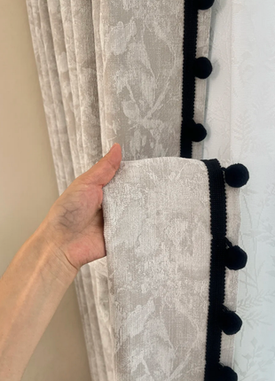 A hand is holding a gray curtain with black trim and pom-poms, which is hanging on a window.