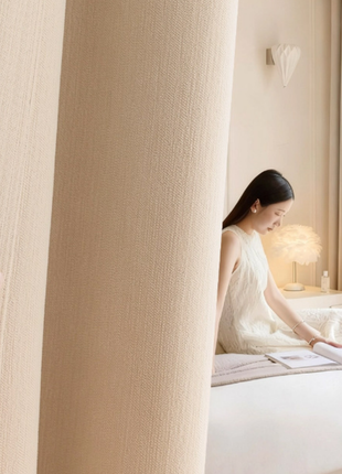 A person is sitting on a bed, reading a book, with a hand holding a curtain in the foreground.