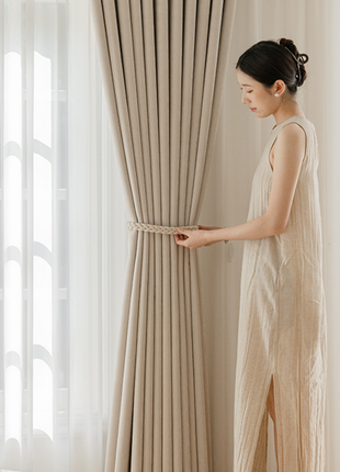 A woman in a long beige dress stands in front of a window, adjusting the beige curtains.