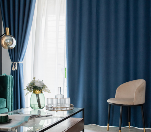 a living room with a blue curtain, a glass coffee table with a vase of flowers and a tray of glasses, a beige armchair, and a white lamp.