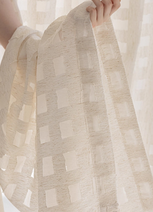 A hand is holding a white fabric with a geometric pattern, partially covering a window.