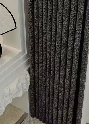 a close-up view of a dark grey curtain with a herringbone pattern, hanging next to a white wall.