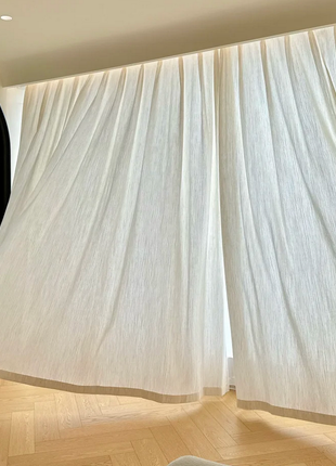 a room with a large window, featuring white curtains that are blowing in the wind. The curtains are open, allowing natural light to enter the room. The wooden floor and the black door frame create a contrast with the light-colored curtains.