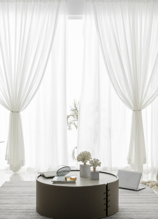 a modern living room with a large window, white curtains, and a round coffee table with a vase of flowers and a book.