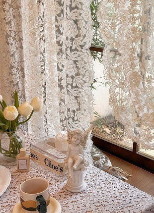 Voilage à broderie florale vu de près devant une fenêtre, tulipes et statuette d'ange sur la table